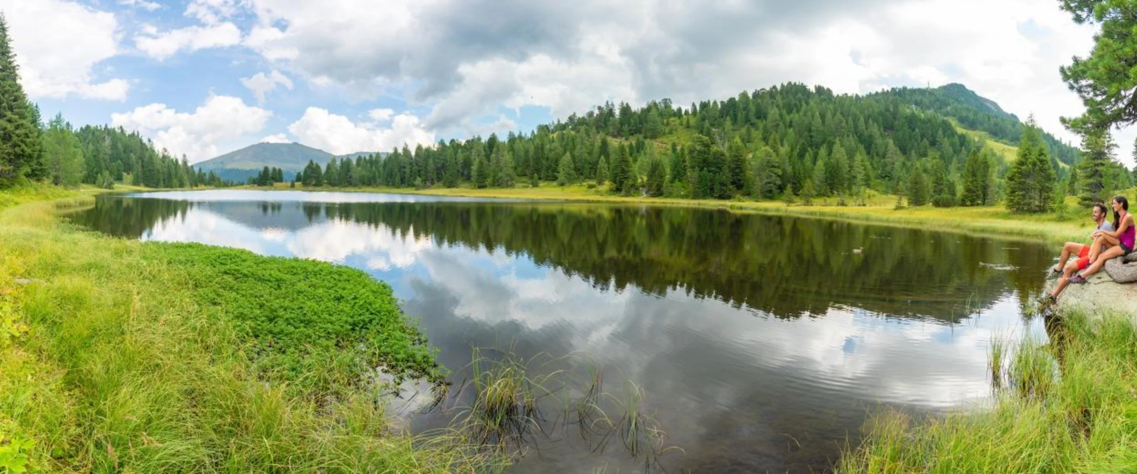 Panoramic view of a serene lake surrounded by lush green forest and hills. Two people sit on rocks near the shore, under a partly cloudy sky.