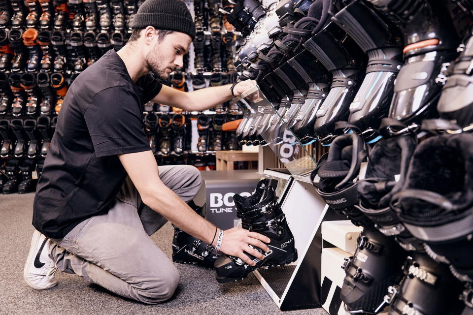 Ein Mann mit Mütze und schwarzem Hemd kniet in einem Skigeschäft und richtet eine Reihe von ausgestellten Skischuhen aus. Der Laden ist mit verschiedenen Skiausrüstungen gefüllt.