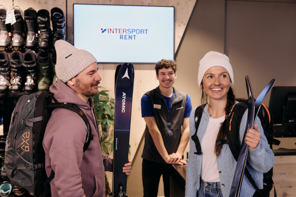 A smiling couple in winter attire holds skis inside a rental shop. A staff member stands behind, under a sign reading "INTERSPORT RENT." The mood is cheerful and welcoming.