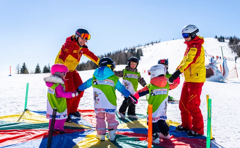 Children in colorful skiing outfits hold hands in a circle on a snowy slope, led by two instructors in red and yellow jackets. A bright, cheerful atmosphere.