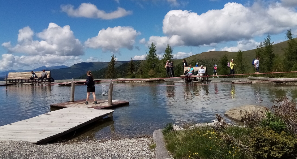 A serene lakeside scene with people enjoying a sunny day, a child on a small dock, lush greenery, fluffy clouds, and distant mountains.