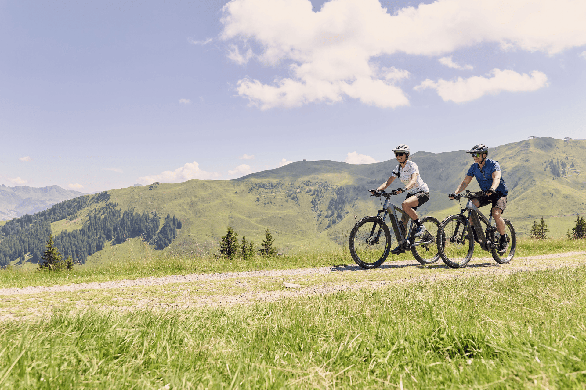 Two people are cycling on a grassy trail with rolling green hills and scattered trees under a clear sky with fluffy clouds, conveying a sense of adventure.