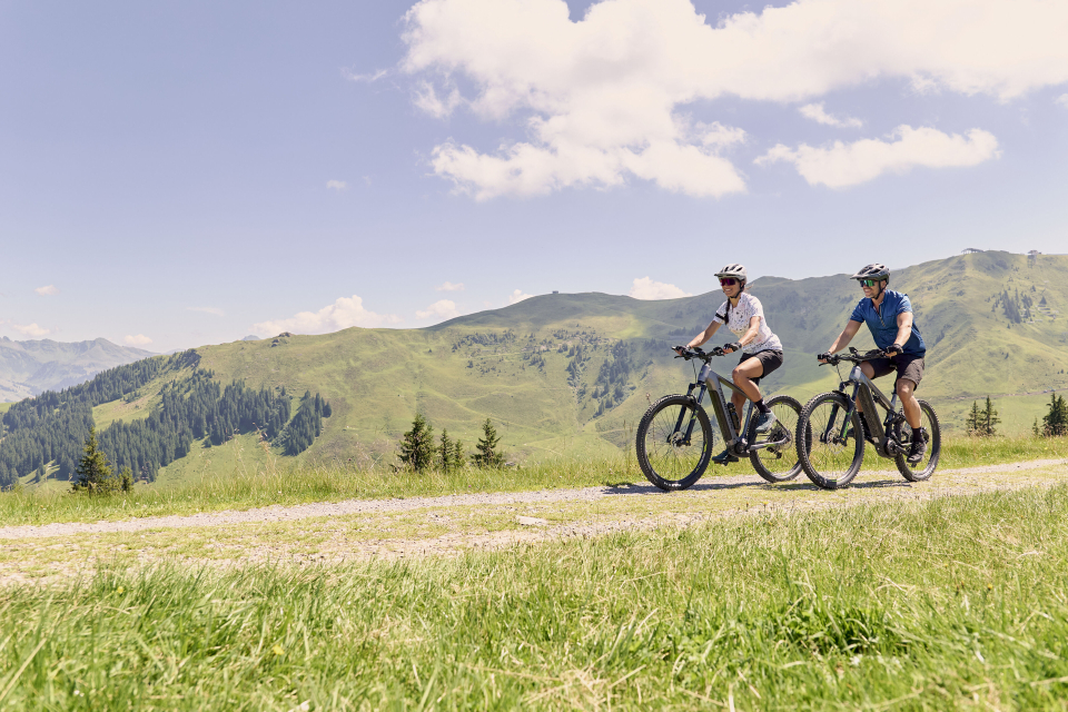 Zwei Menschen radeln auf einem grasbewachsenen Weg mit sanften grünen Hügeln und vereinzelten Bäumen unter einem klaren Himmel mit flauschigen Wolken und vermitteln dabei ein Gefühl von Abenteuer.