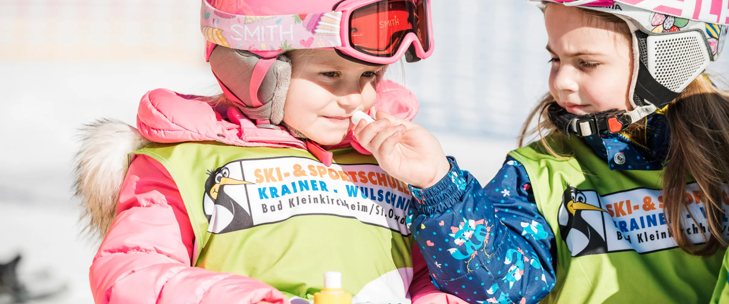 Two young children wearing colorful ski gear and helmets, sharing a playful moment on the snowy slopes. They are smiling and appear cheerful.