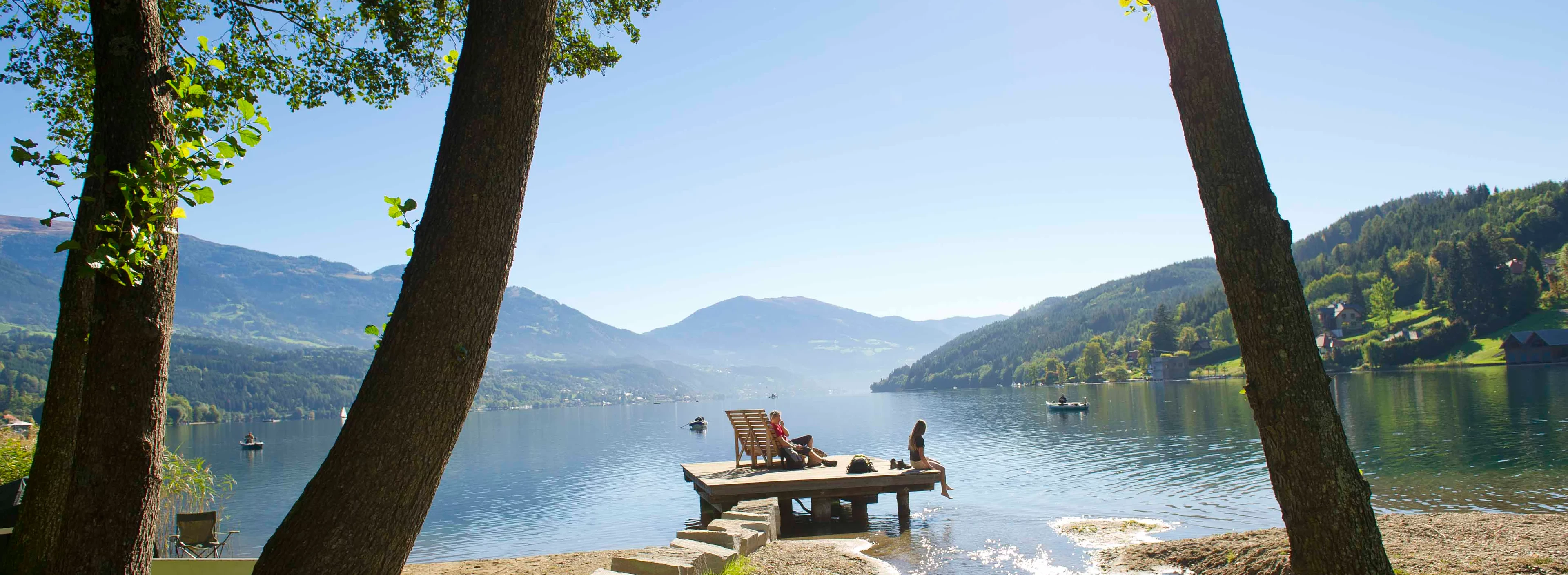 A serene lakeside scene with a wooden dock extending into calm water. Two people relax on the dock. Framing trees and distant hills under a clear sky.