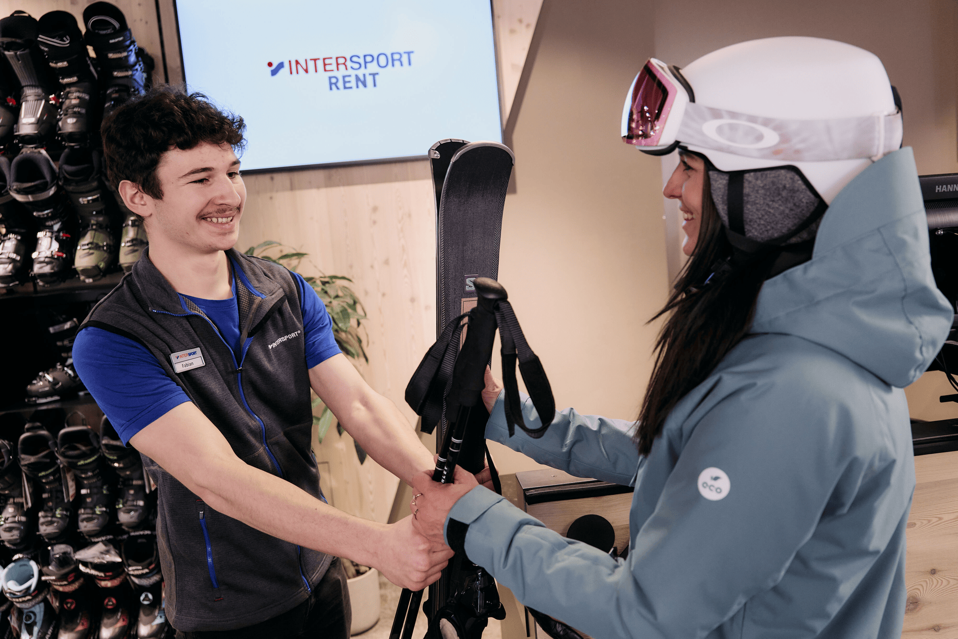 A smiling store associate hands skis to a customer in winter gear at a ski rental shop. Shelves with ski boots and a screen with "INTERSPORT RENT" are visible.