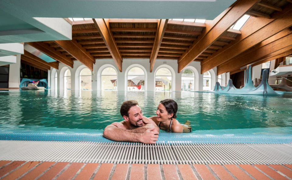 Ein Paar lächelt sich in einem Innenpool mit Holzbalken und Bogenfenstern an. Die Umgebung ist ruhig und einladend und vermittelt Entspannung.