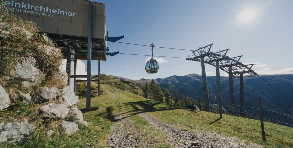 Cable car at a mountain station, suspended above a green landscape under a clear blue sky. Rugged terrain and distant peaks create a serene atmosphere.