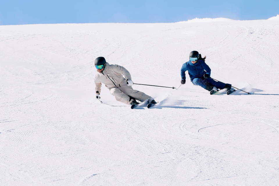 Two skiers in winter gear carve through pristine snow on a sunny day. The skier in gray leads, angled sharply, with another in blue closely following.