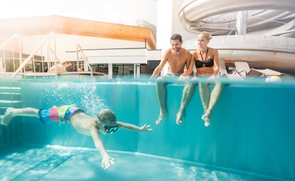 Ein Junge schwimmt mit einer Schwimmbrille unter Wasser in einem Pool. Ein Mann und eine Frau sitzen am Beckenrand und lächeln. Im Hintergrund ist eine große Wasserrutsche zu sehen. Eine helle und lebhafte Szene.