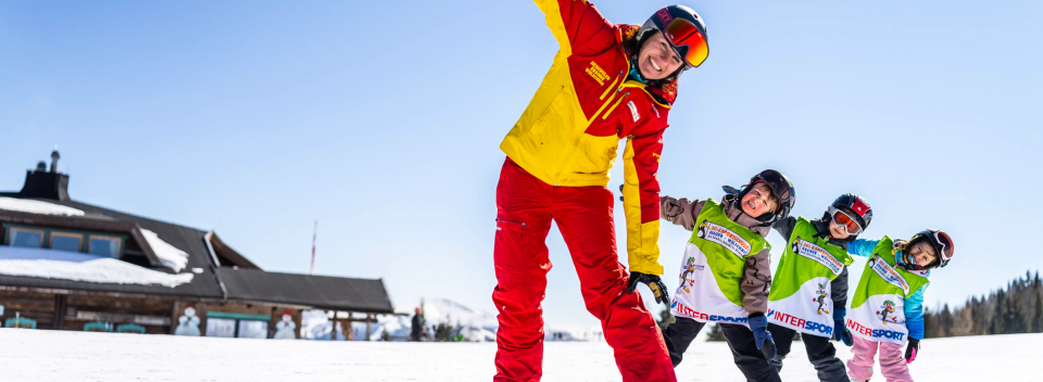 Ski instructor in red and yellow leads three smiling children in a fun ski lesson on a sunny, snowy slope. A lodge is visible in the background.