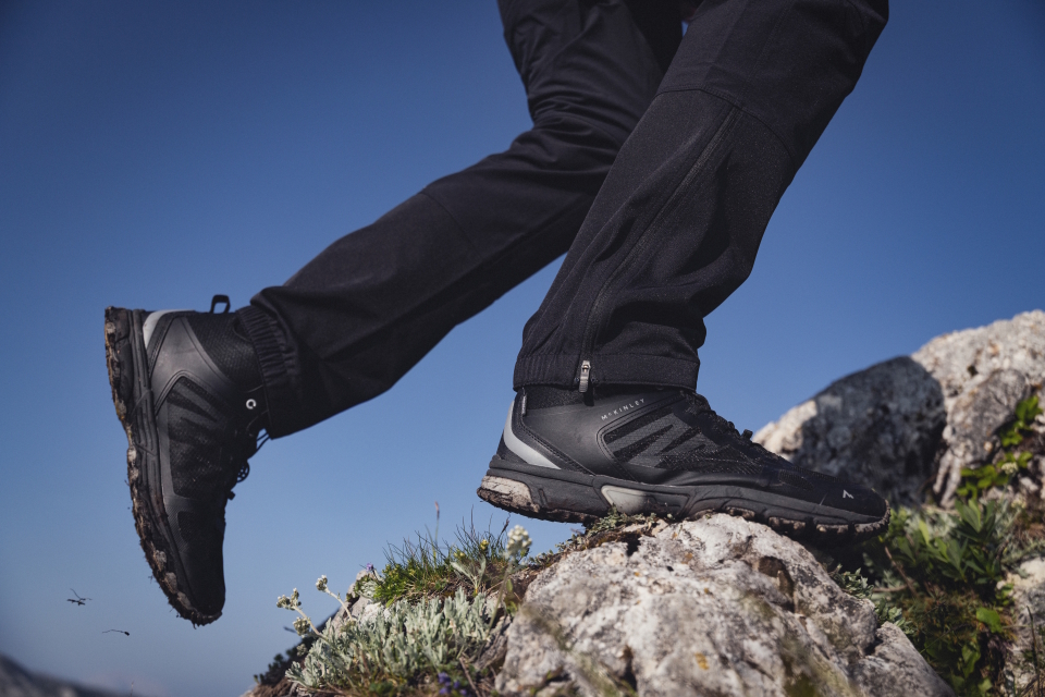 Black hiking shoes stepping on rocky terrain with alpine plants under a clear blue sky.