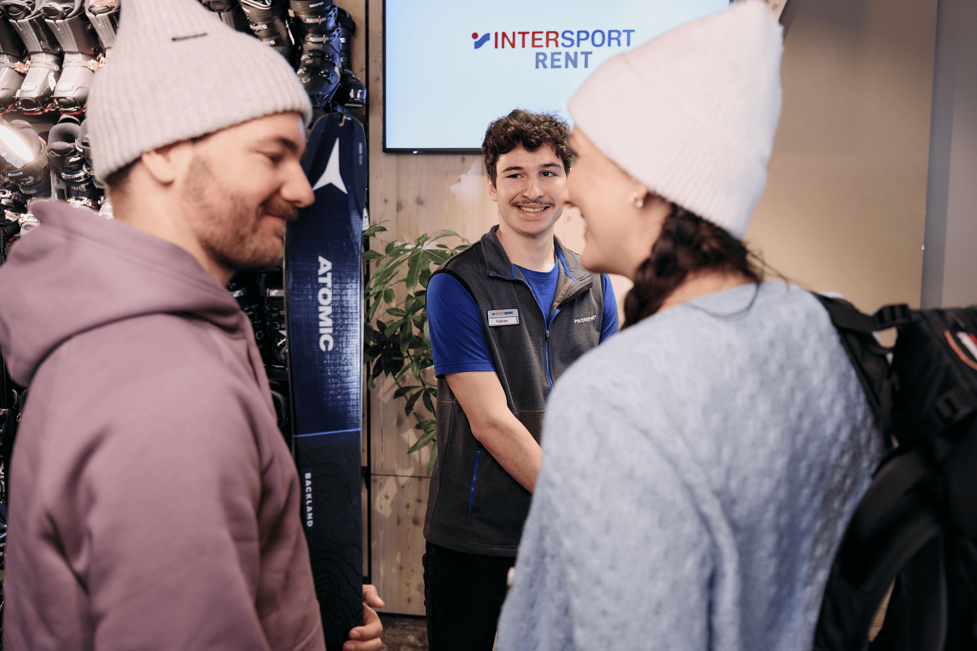 A smiling store employee assists a man and woman in winter clothing at a ski rental shop. The mood is cheerful and welcoming, with rental gear visible.