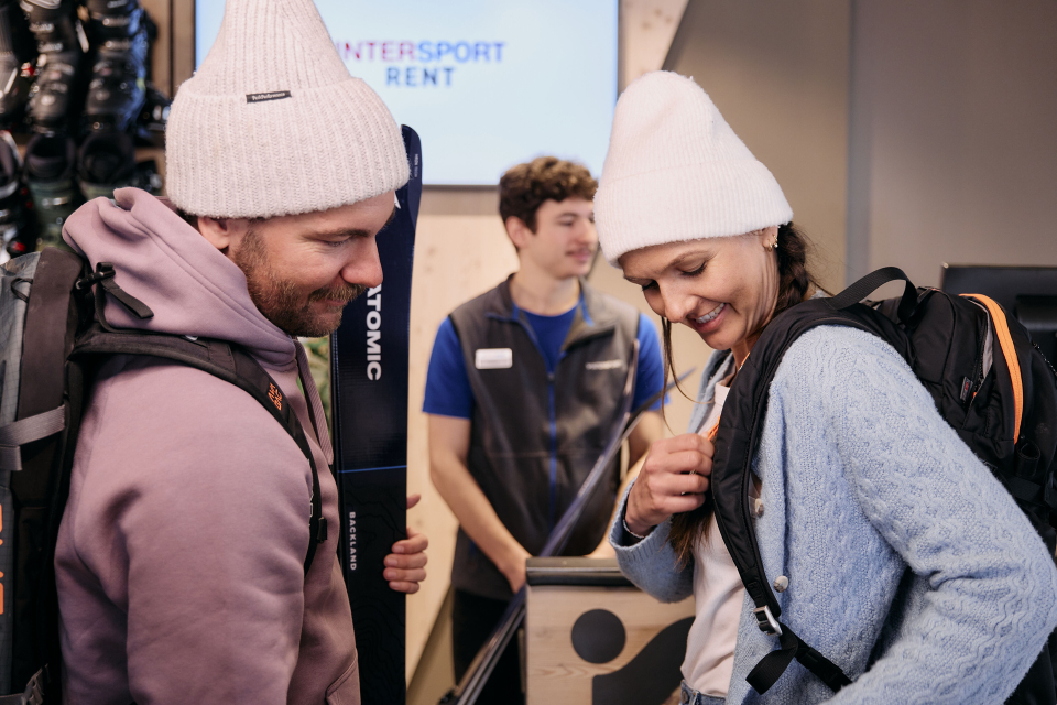 A man and woman, both wearing white beanies, smile while adjusting their backpacks in a ski rental shop. A staff member assists in the background.