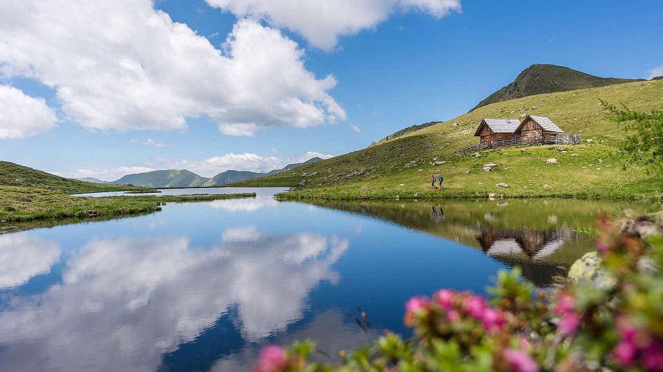 Eine ruhige Berglandschaft mit einem klaren See, in dem sich die Wolken spiegeln. Eine rustikale Hütte steht auf einem grasbewachsenen Hügel, im Vordergrund leuchtend rosa Blumen.