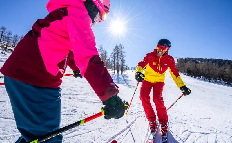 Two skiers, one in red and yellow, the other in pink and blue, glide down a snowy slope under a clear blue sky and bright sun, conveying joy and excitement.