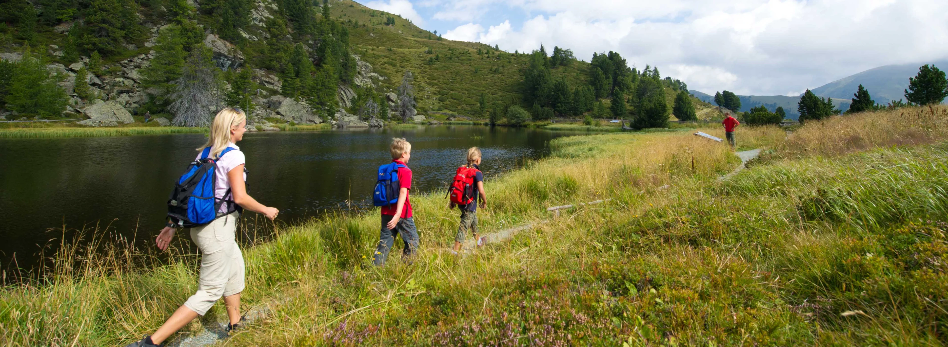 A family hikes on a grassy trail beside a calm lake and forested hills under a partly cloudy sky, conveying a sense of adventure and tranquility.