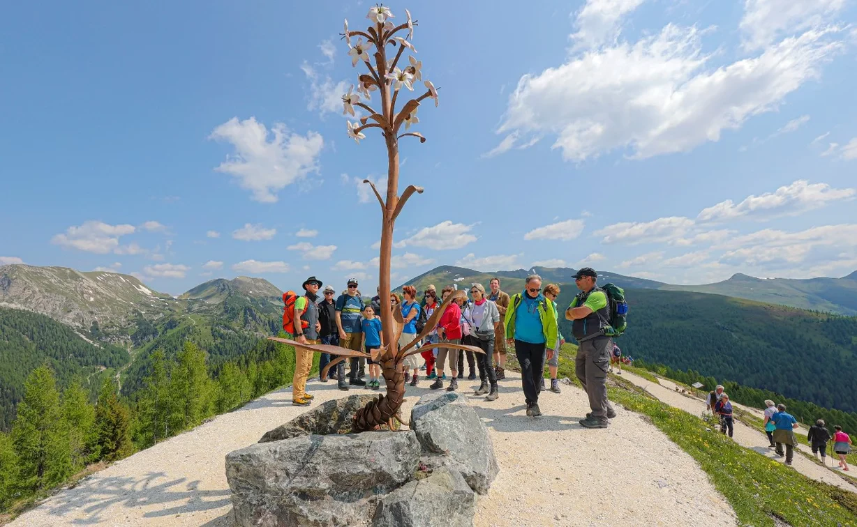 Group of hikers in vibrant clothing admire a metal sculpture on a mountain path. Blue sky with clouds, green landscape, and a cheerful atmosphere.
