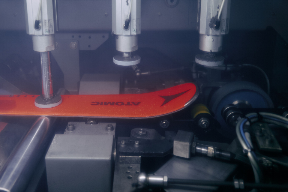 A ski being precisely manufactured with automated machinery. The orange ski reads "Atomic" and is surrounded by various mechanical components in a factory setting.