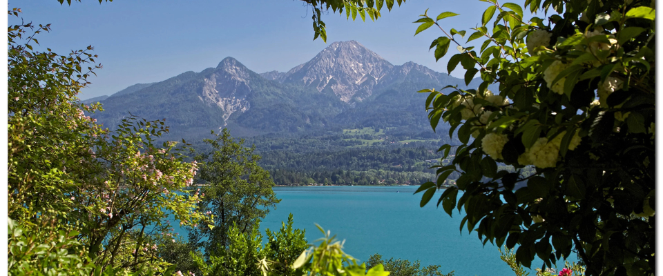 Ein ruhiger Blick auf einen türkisfarbenen See, umrahmt von üppigem Grün, mit majestätischen Bergen im Hintergrund unter einem klaren blauen Himmel. Friedlich und malerisch.