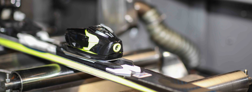 Nahaufnahme einer Skibindung, die in einer Werkstatt gewartet wird, mit Fokus auf das schlanke, moderne Design und die Präzisionsmaschinen. Der Ton ist technisch und professionell.