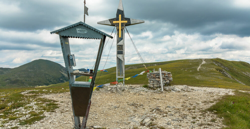 A weathered metal sign and a tall cross adorned with prayer flags stand on a rocky hilltop under a cloudy sky, conveying a sense of serenity and remoteness.