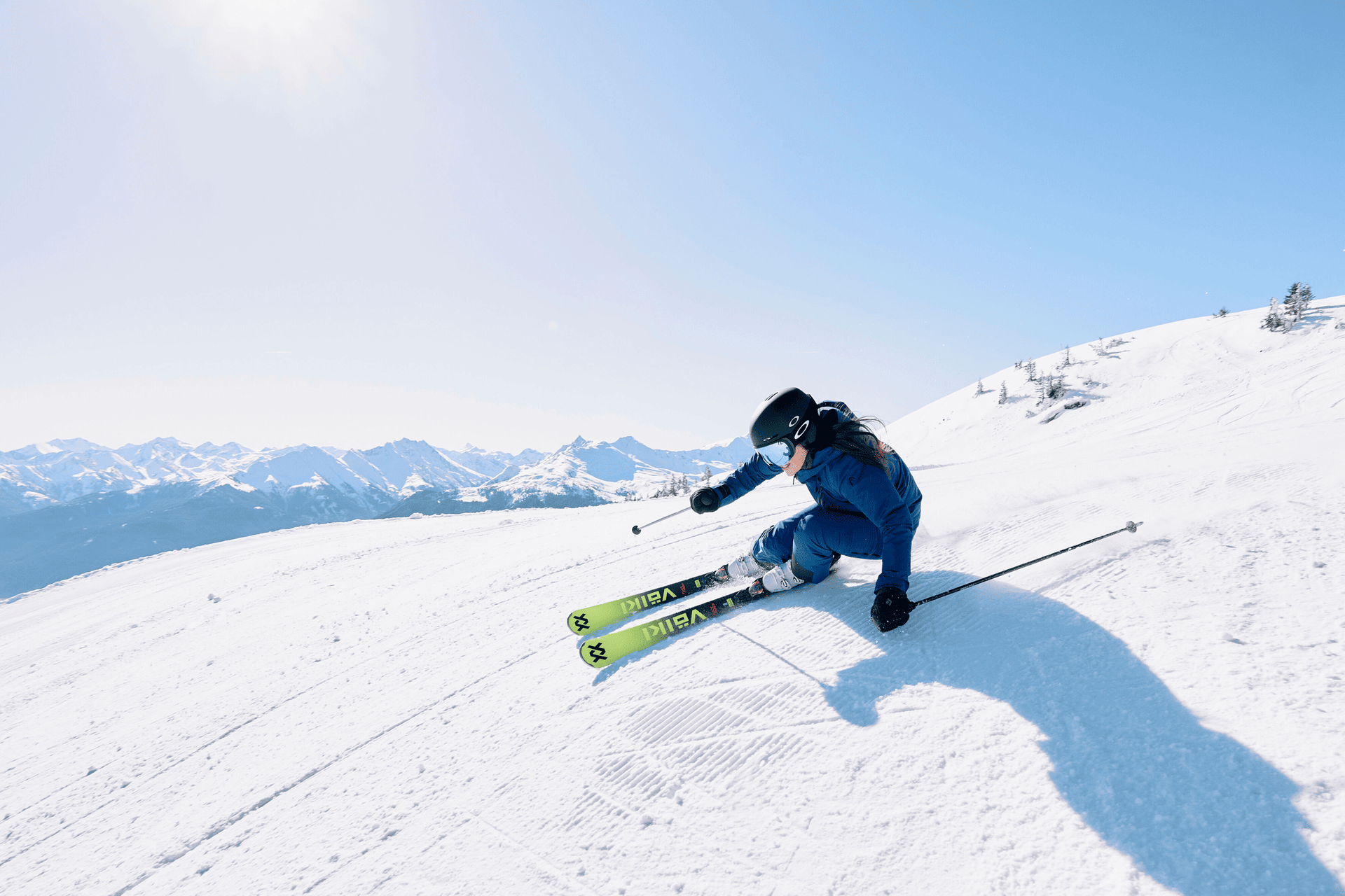 A skier in blue gear expertly navigates a snowy slope under a clear sky. Sunlight casts sharp shadows and highlights the distant snow-capped mountains.