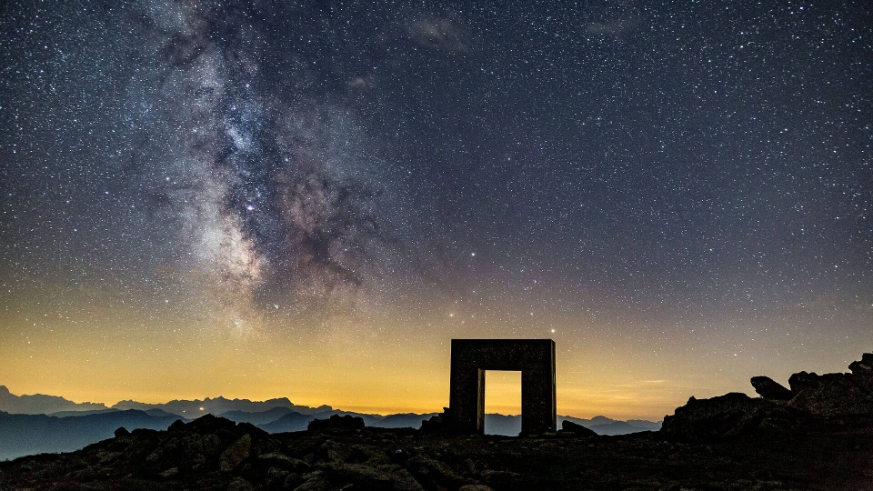 Starry night sky with the Milky Way above a silhouetted stone archway on rocky terrain. A warm glow on the horizon suggests a serene, mysterious atmosphere.