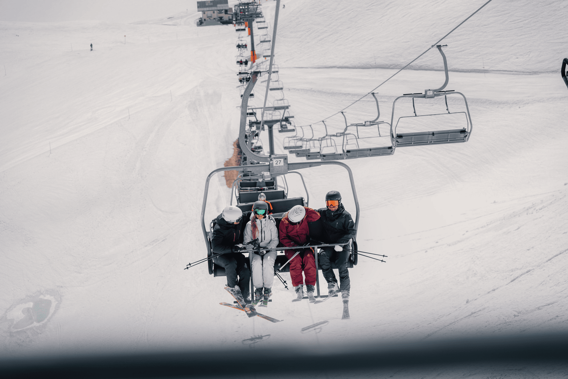 Four skiers ride a snowy ski lift, bundled in winter gear. They are seated close together, creating a sense of camaraderie amidst the vast, white landscape.