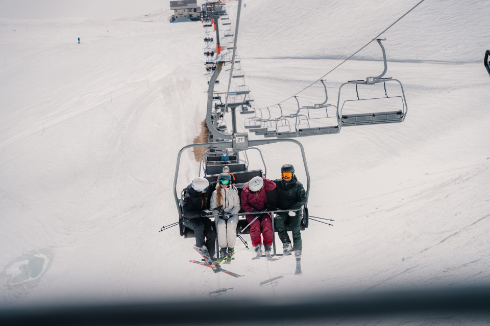 Vier Skifahrer fahren in Winterkleidung auf einem verschneiten Skilift. Sie sitzen eng beieinander und schaffen so inmitten der weiten, weißen Landschaft ein Gefühl der Kameradschaft.