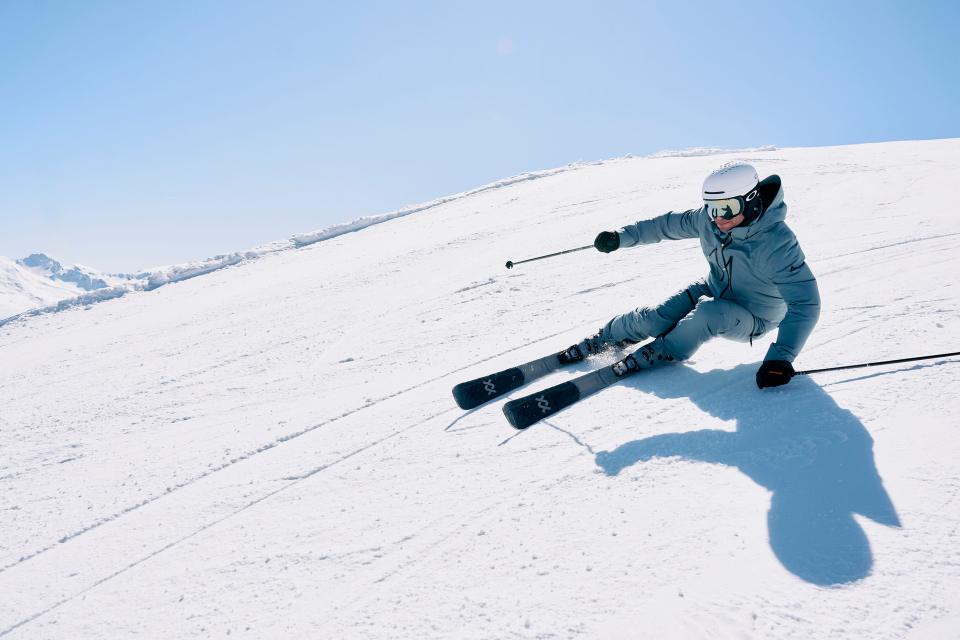 Skier in a blue outfit expertly carving down a snowy slope under a clear blue sky. Shadows accentuate the dynamic movement and skill.