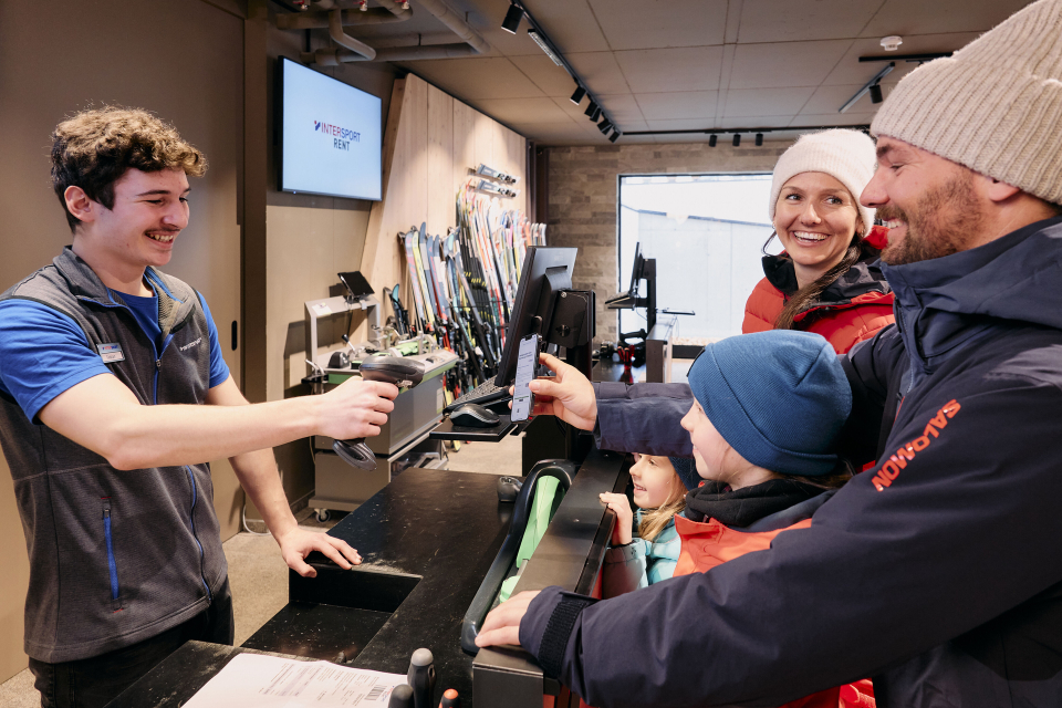 A smiling store clerk scans a phone held by a father, with a family dressed in winter clothing. Ski equipment is displayed in the background.