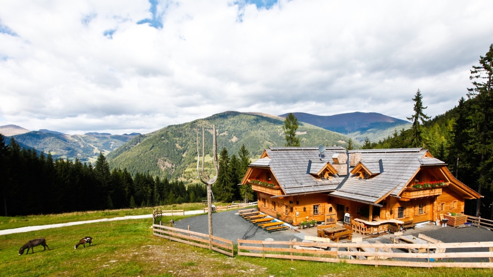 A cozy wooden cabin sits in a lush mountain landscape under a partly cloudy sky. Grazing deer in the foreground and vast forests evoke tranquility.