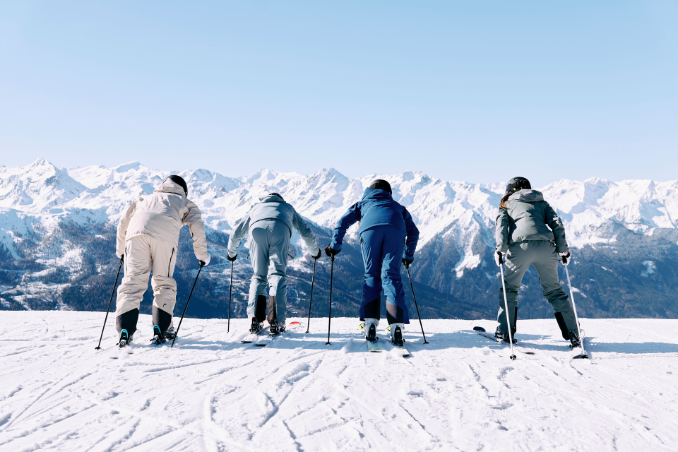 Four skiers in winter gear stand at the snow-covered hilltop, poised to descend. Snowy mountains stretch in the background under a clear sky.