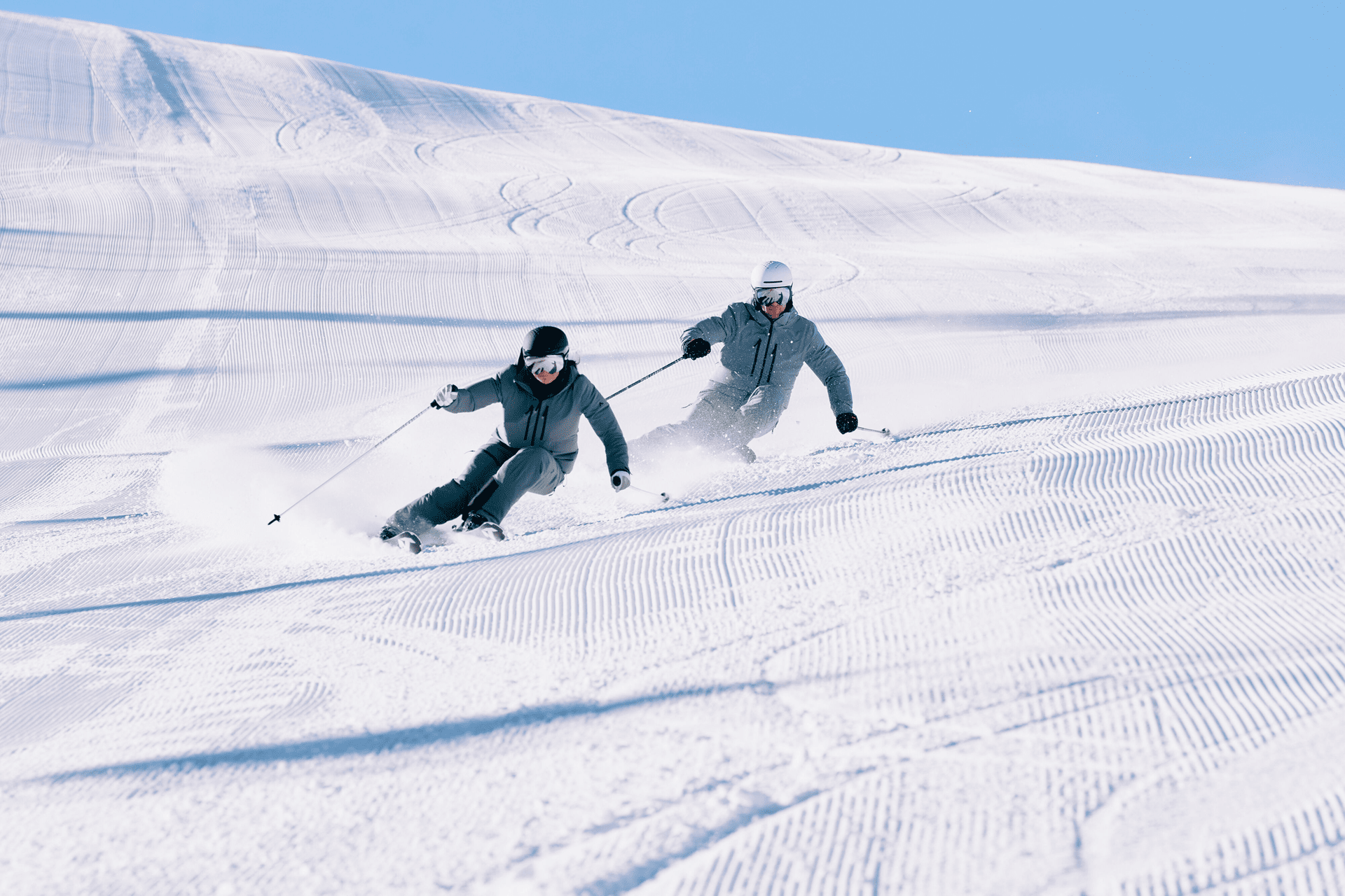 Zwei Skifahrer in blauer Ausrüstung gleiten schnell einen sonnenbeschienenen, präparierten Hang hinunter und hinterlassen Spuren im Schnee. Der klare Himmel unterstreicht die berauschende Szenerie.