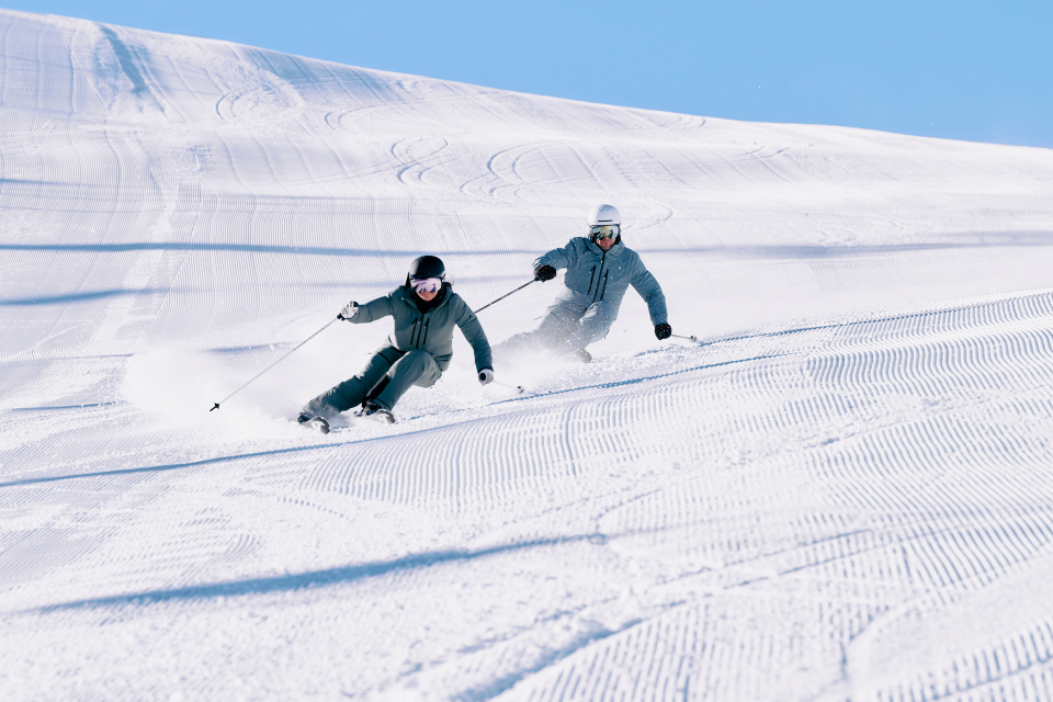Two skiers in blue gear glide swiftly down a sunlit, groomed slope, leaving trails in the snow. The clear sky enhances the exhilarating scene.
