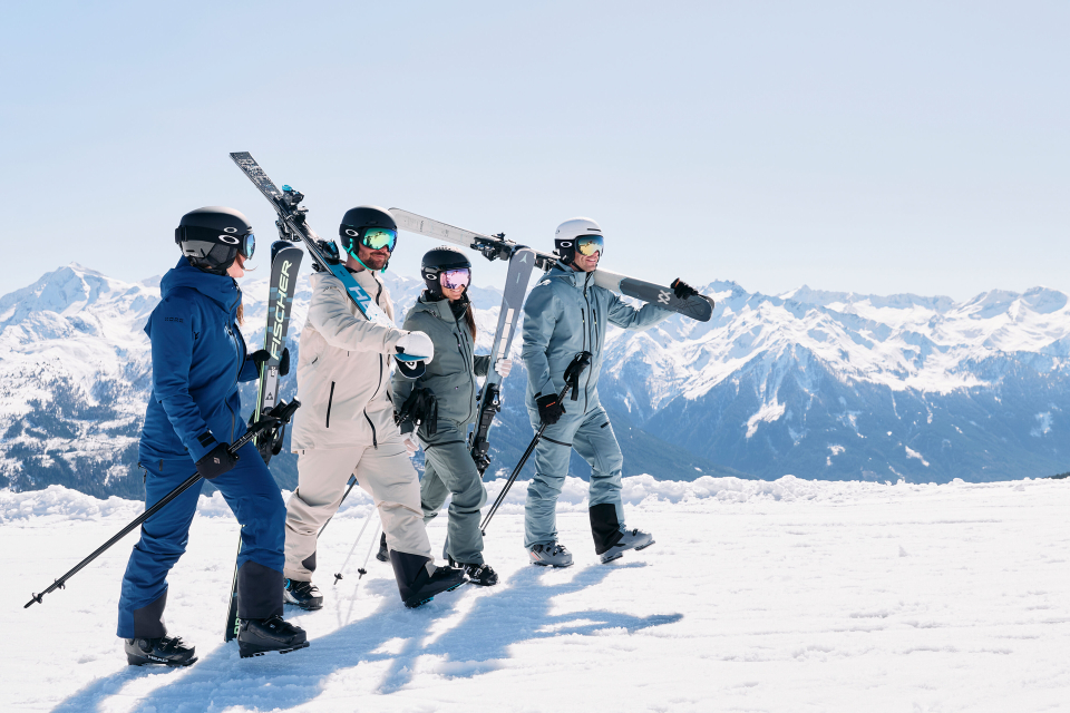 Four people in ski gear walk across a snowy mountain, with one carrying skis over their shoulder. Bright, clear skies and majestic mountains in the background convey a sense of adventure and camaraderie.