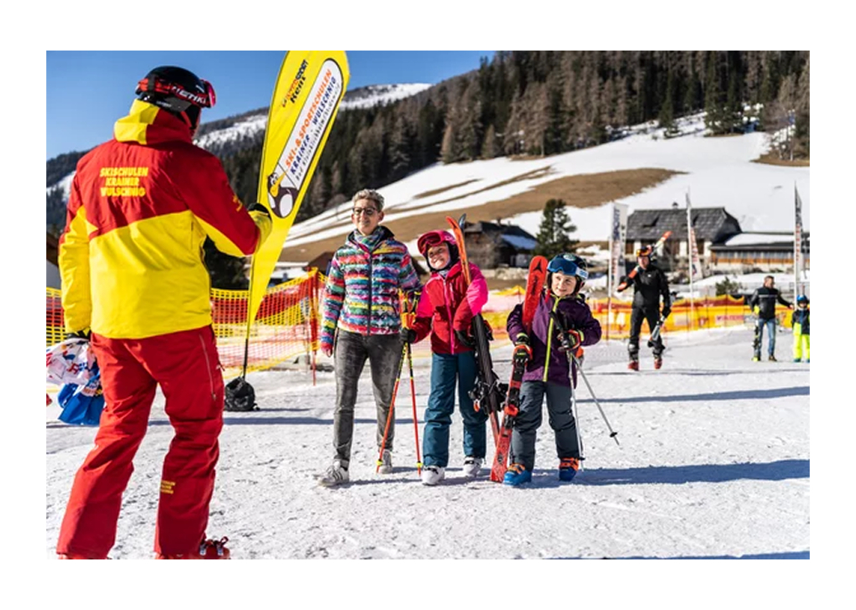 Ein Kind in bunter Skikleidung und ein Pinguin-Maskottchen fahren spielerisch auf Skiern einen verschneiten Hang hinunter unter einem strahlend blauen Himmel, im Hintergrund eine Berghütte.