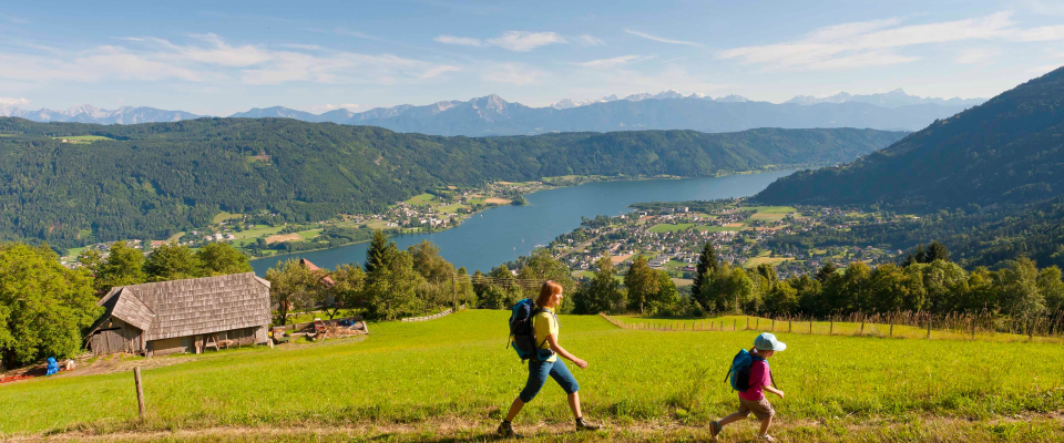 Two hikers are walking on a grassy hill overlooking a picturesque valley with a lake, mountains, and a village. The scene is bright and peaceful.