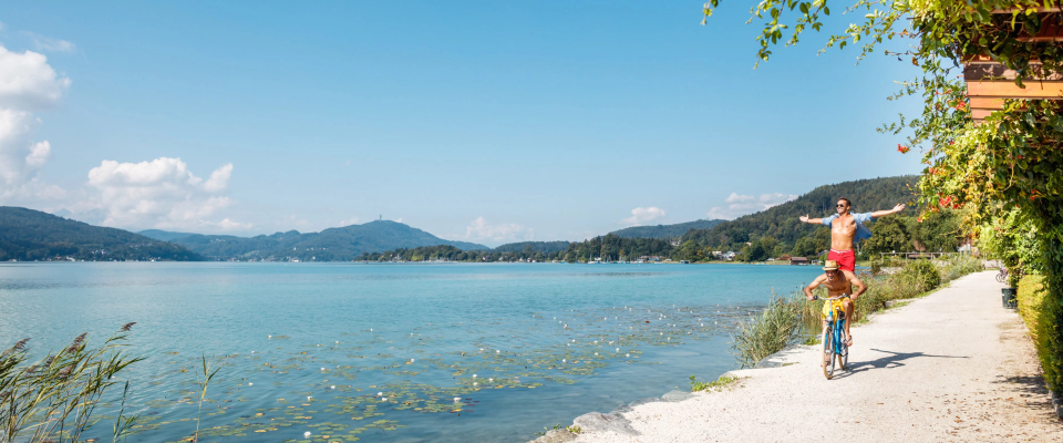 Ein Vater balanciert fröhlich auf den Schultern seines Kindes, während er an einem ruhigen, klaren blauen See entlangradelt. Berge und Grünflächen bilden eine friedliche Sommerkulisse.