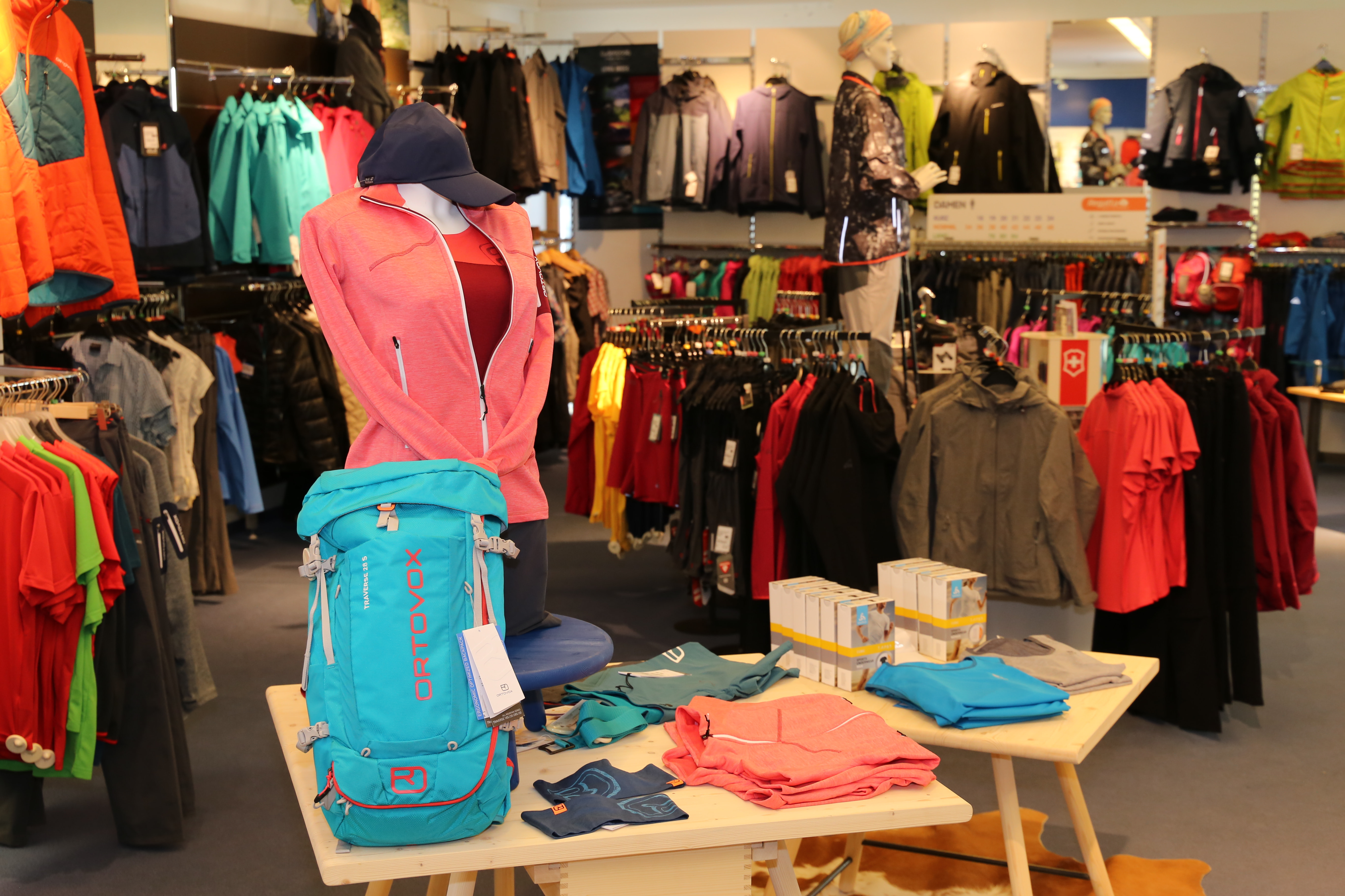 Ein lebhaftes Outdoor-Bekleidungsgeschäft präsentiert farbenfrohe Jacken, Hemden, Rucksäcke und Wanderausrüstung. Eine Schaufensterpuppe trägt eine rosa Jacke und vermittelt damit einen abenteuerlichen Eindruck.