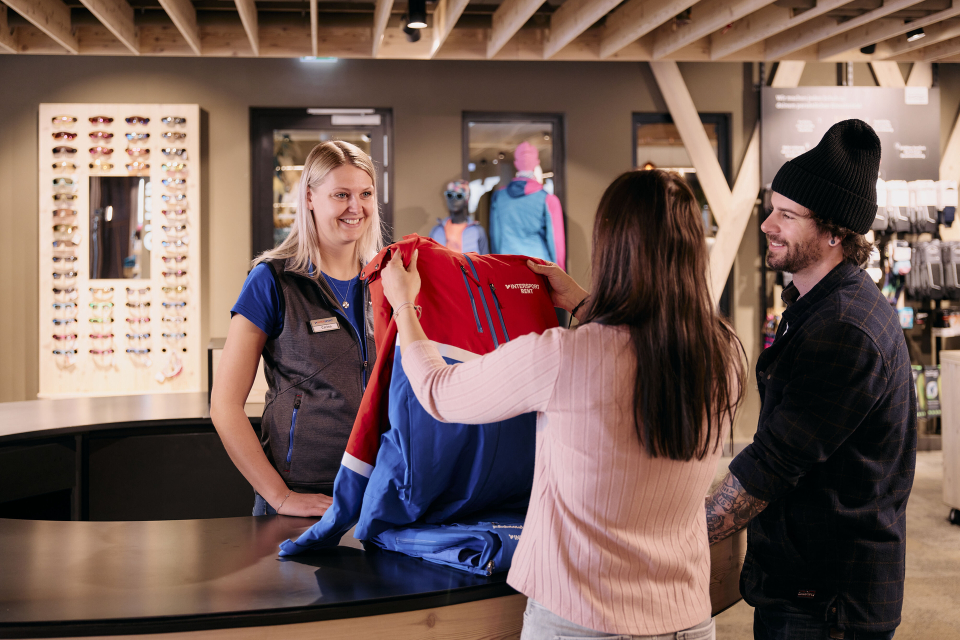 Ein lächelnder Verkäufer bedient eine Frau und einen Mann an einer Theke mit einer roten und blauen Jacke. Der Schauplatz ist ein gut beleuchteter, einladender Laden mit ausgestellten Waren.