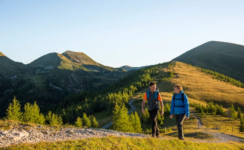 Zwei Wanderer mit Rucksäcken wandern unter einem strahlend blauen Himmel auf einem grasbewachsenen Bergpfad. Das Sonnenlicht wirft sanfte Schatten auf die sanften Hügel und das üppige Grün.