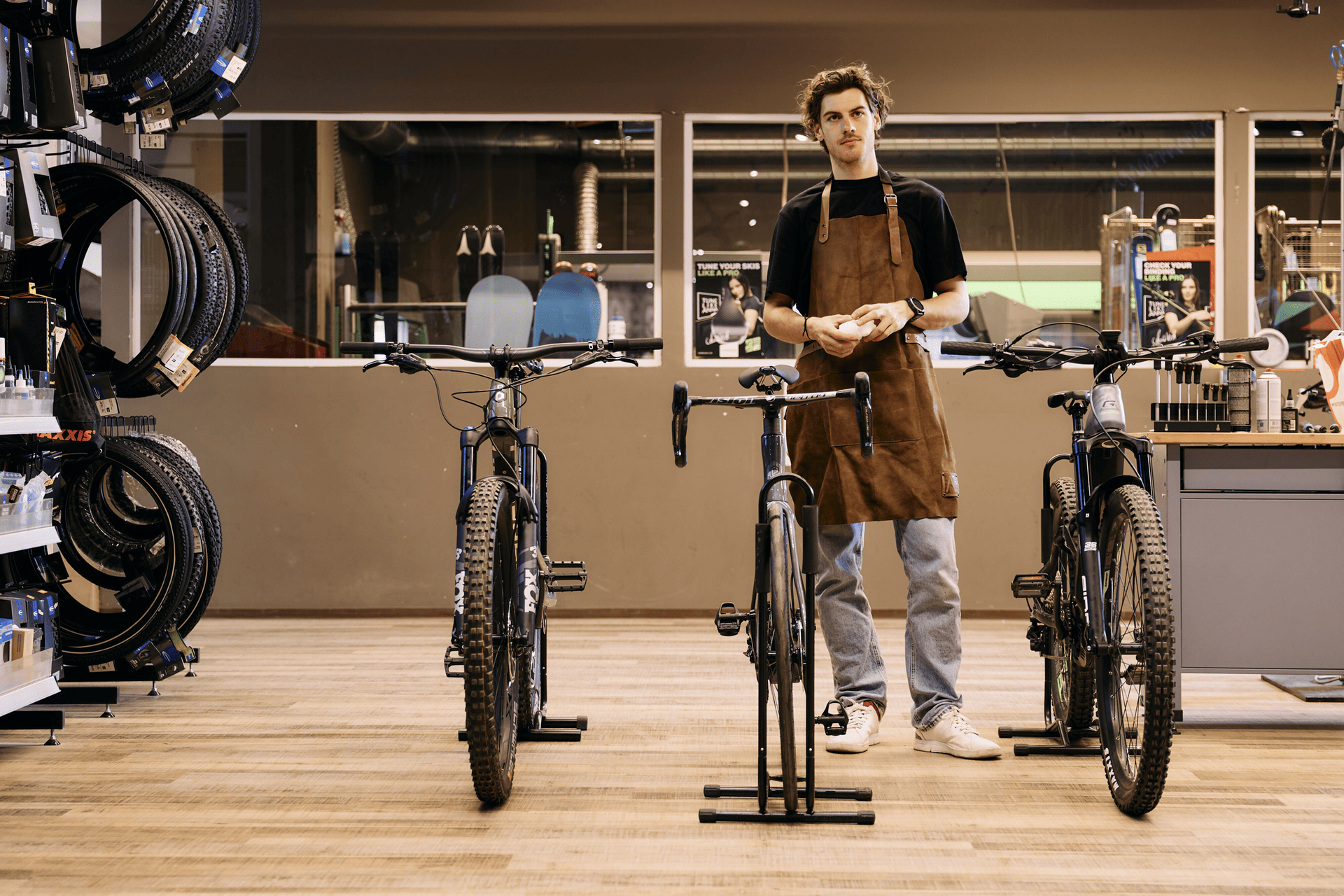 Young man in apron stands confidently in a bike shop, flanked by three bicycles. Shelves with tires are on the left, creating a focused, industrious atmosphere.