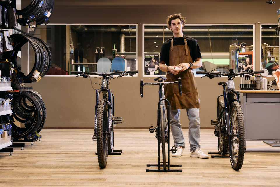 Ein junger Mann in einer Schürze steht selbstbewusst in einem Fahrradgeschäft, flankiert von drei Fahrrädern. Links befinden sich Regale mit Reifen, die eine konzentrierte, fleißige Atmosphäre schaffen.