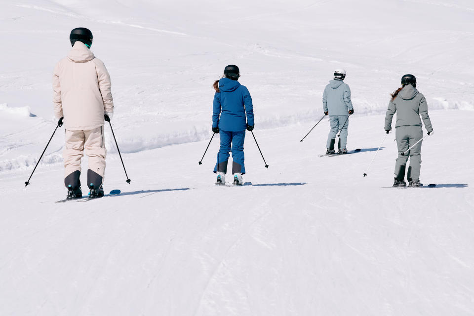 Vier Skifahrer in bunten Outfits fahren mit dem Rücken zum Betrachter einen verschneiten Hang hinunter, unter einem klaren Himmel. Die Szene vermittelt ein Gefühl von Abenteuer und Ruhe.