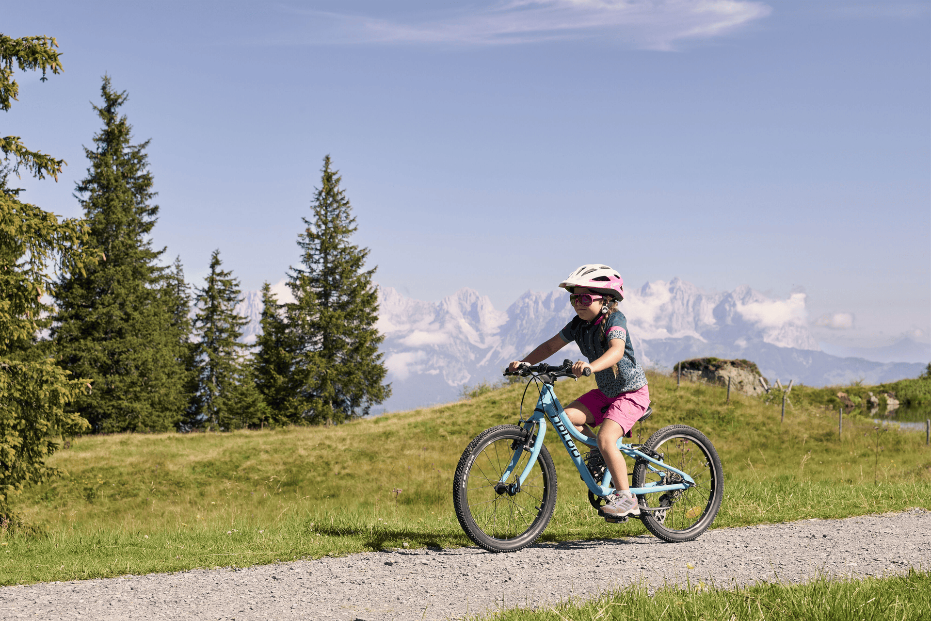 A child in a pink helmet rides a blue bicycle on a gravel path surrounded by green grass and trees, with majestic mountains in the background.
