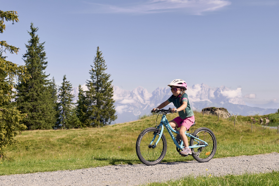 Ein Kind mit einem rosa Helm fährt auf einem blauen Fahrrad auf einem Schotterweg, umgeben von grünem Gras und Bäumen, mit majestätischen Bergen im Hintergrund.
