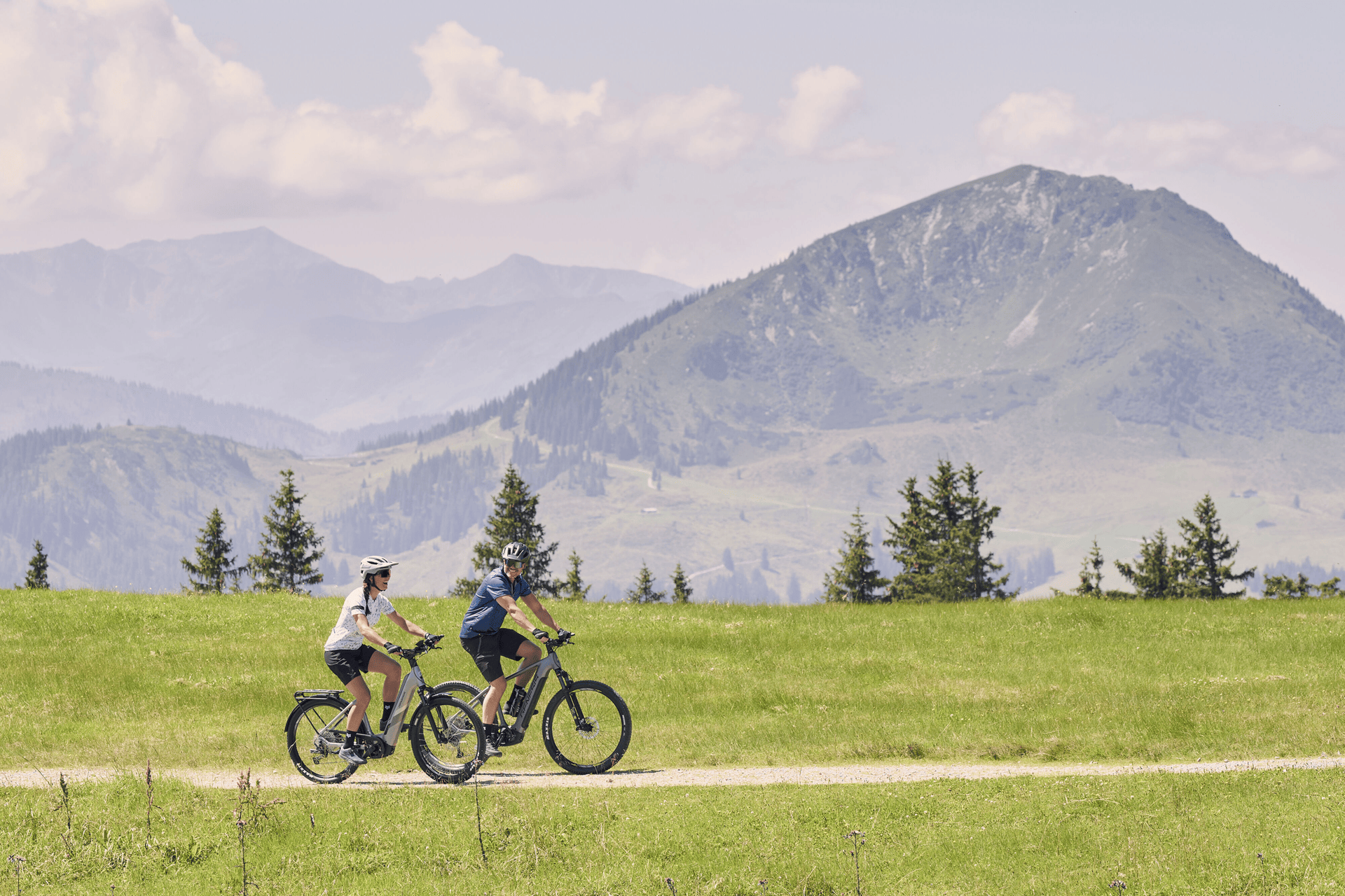 Two people ride bikes on a grassy trail with mountains in the background. The scene is calm and sunny, with a clear sky and distant trees.