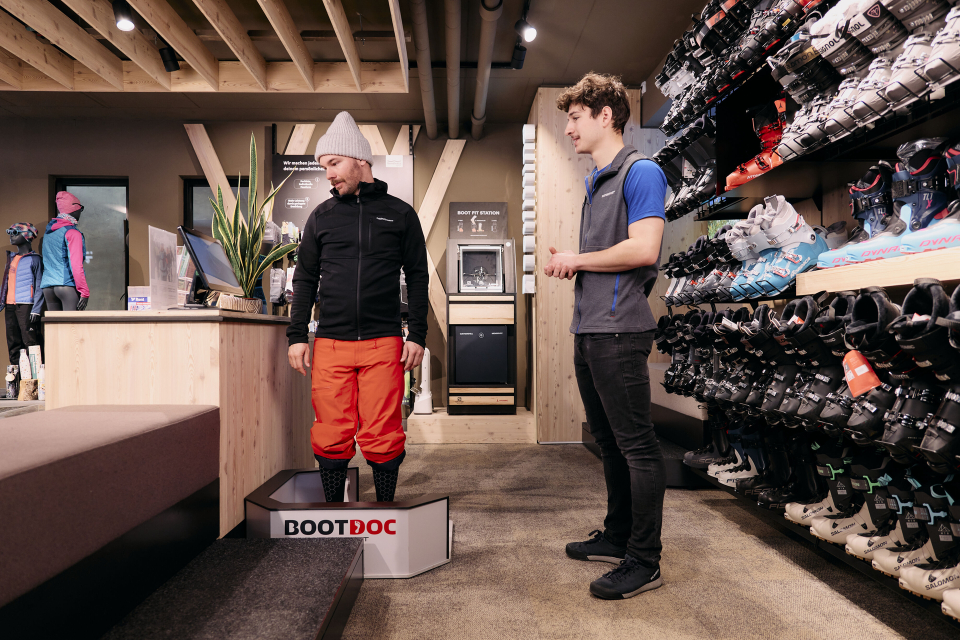 Two people are in a ski shop. One is standing on a fitting platform wearing orange ski pants, and the other is assisting. Shelves of ski boots line the wall.